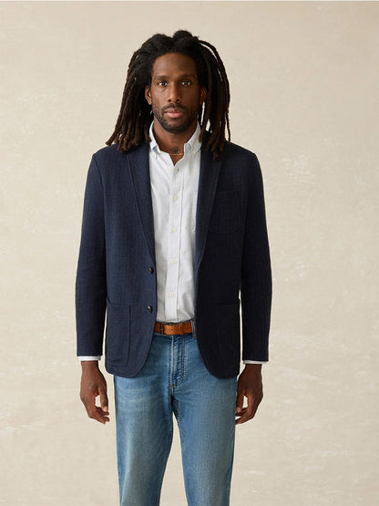 A person with long hair, dressed in the comfortable Faherty Brand Inlet Herringbone Blazer in Navy Twilight, paired with a white shirt and jeans, poses against a neutral background.