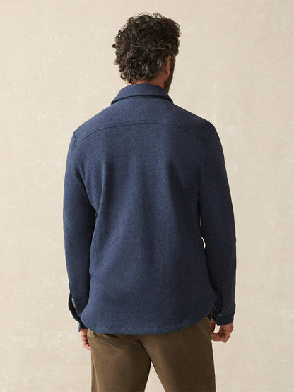 A man with curly hair, seen from behind, wears the Faherty Brand Inlet Knit CPO in Navy Night Melange and brown pants against a plain light background.