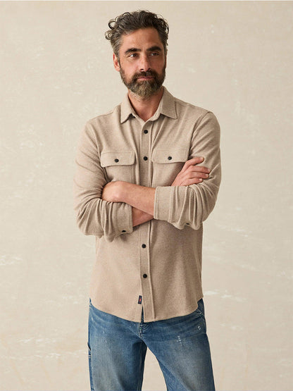 A bearded man in the Faherty Brand Legend Sweater Shirt in Timber Twill and jeans stands with arms crossed against a neutral background, embodying Faherty's rugged style.