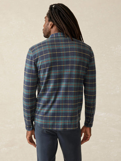 A person with long hair, dressed in the Mossy Elm Plaid Legend Sweater Shirt by Faherty Brand, stands with dark pants, facing away.