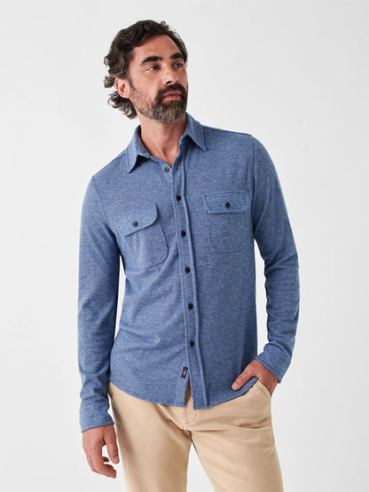 A man wearing the Faherty Brand Legend Sweater Shirt in Glacier Blue Twill and beige pants poses against a plain background, highlighting a versatile layering piece that adds comfort to any outfit.