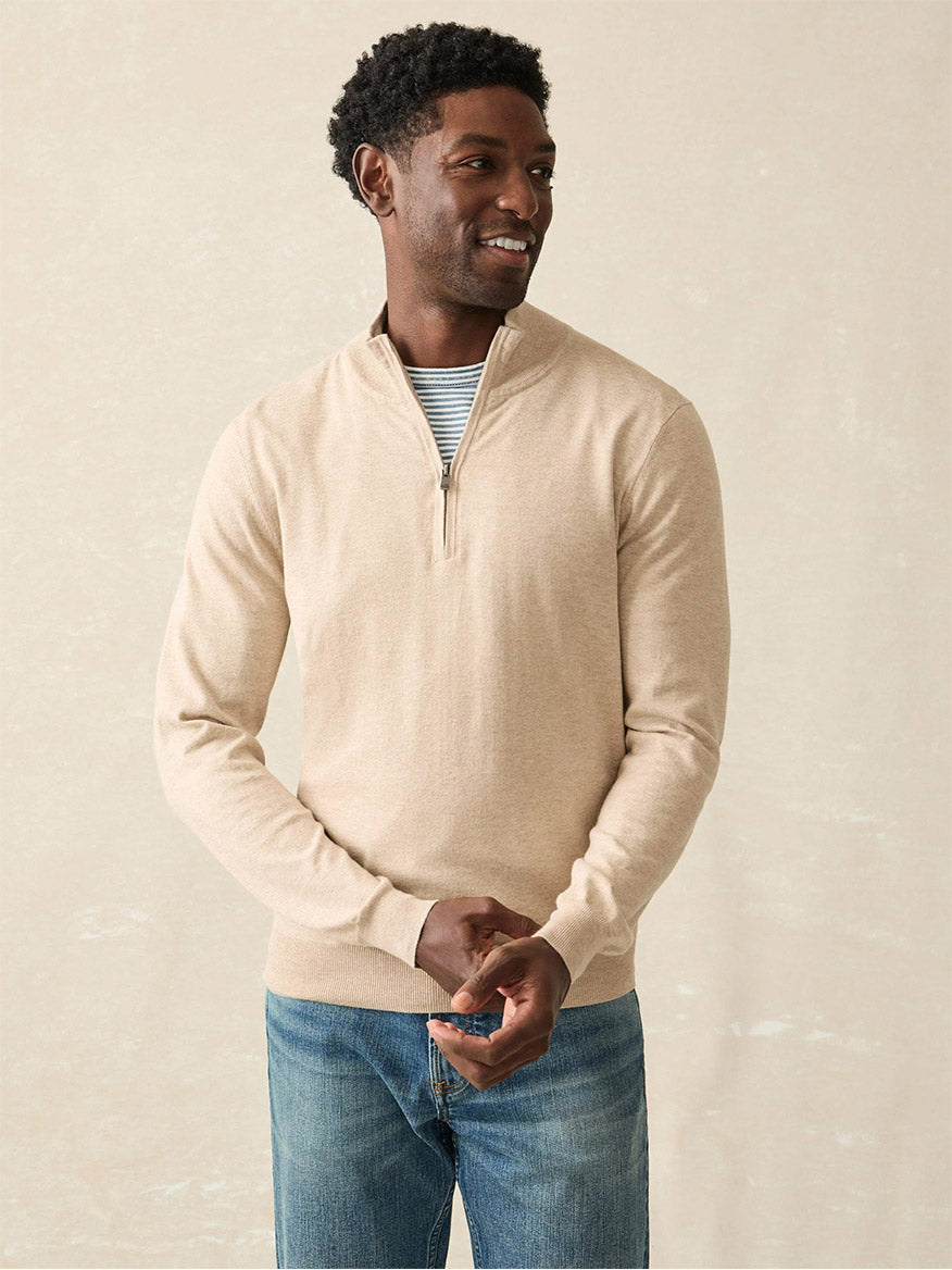 A person wearing the Faherty Brand Movement Quarter Zip in Soft Dune Heather, featuring a striped shirt underneath and crafted from COOLMAX blend performance fabric, stands against a neutral background.