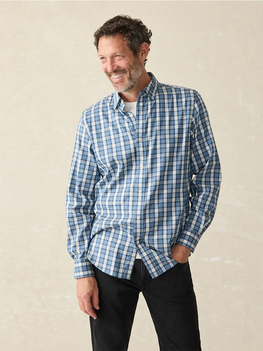 A man wearing the Faherty Brand Movement Shirt in Seaside Blue Plaid and black pants stands with one hand in his pocket, smiling.