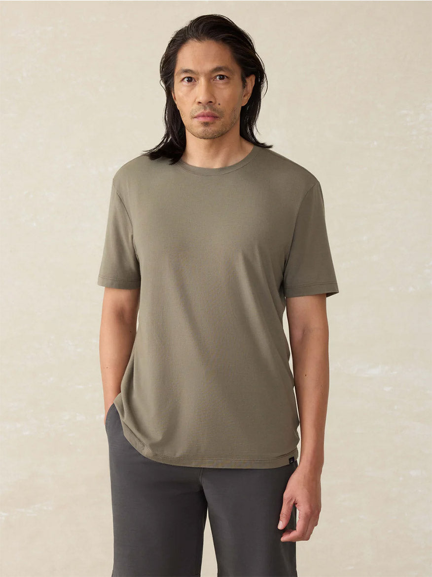 A man with long dark hair wears the Movement Short-Sleeve T-Shirt in Olive Pine Feeder and dark pants, standing against a light beige background.