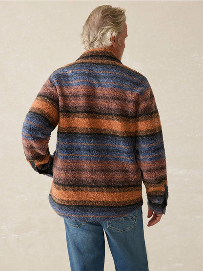 A person with light hair is standing, showing their back. They are wearing the Faherty Brand Range Fleece CPO in Autumn Horizon Stripe, which features rugged shades of orange, brown, and blue. The jacket's recycled high-pile fleece guarantees warmth and comfort.