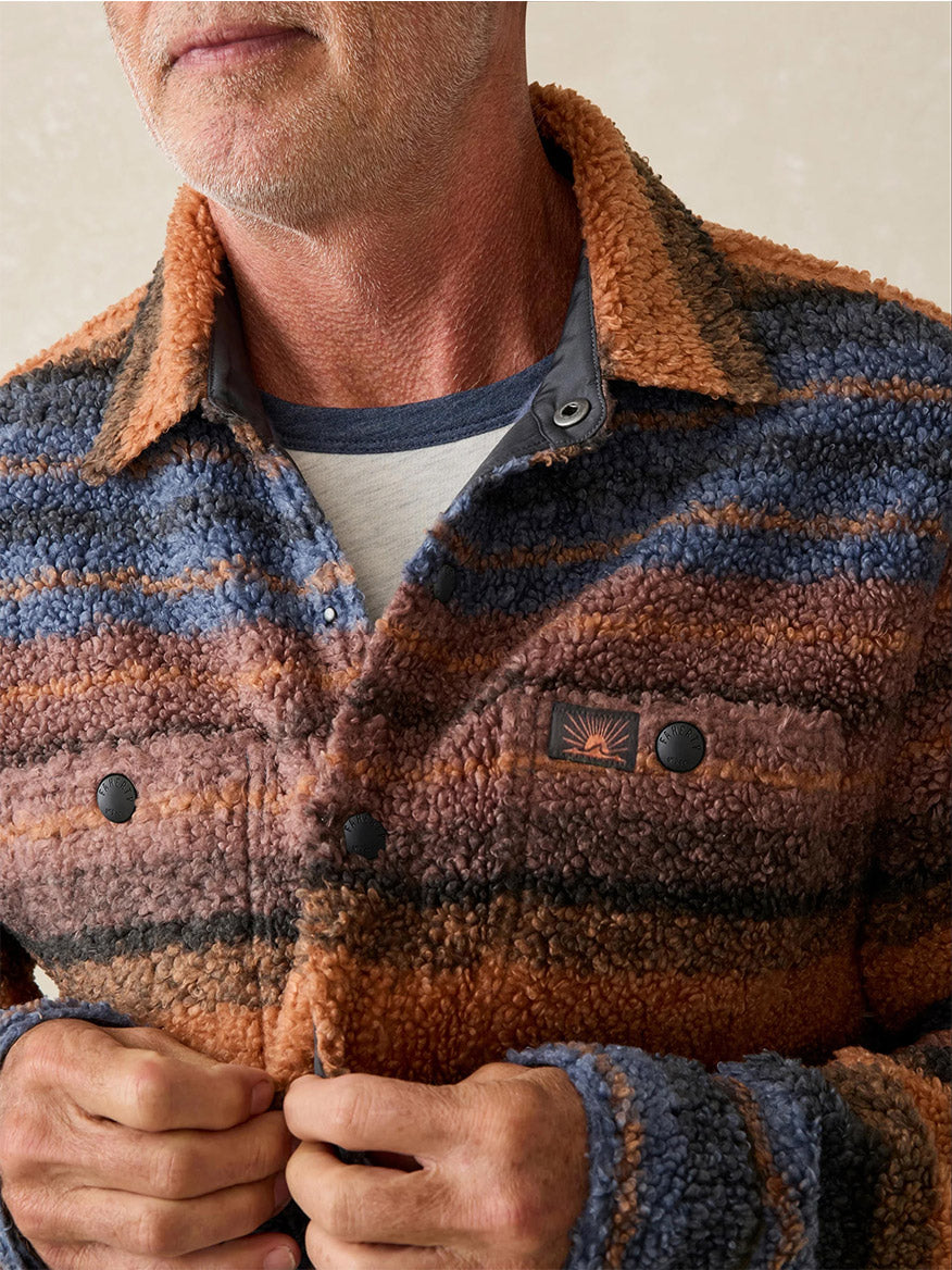 A person wearing the Faherty Brand Range Fleece CPO in Autumn Horizon Stripe, buttoned over a gray and blue shirt, showcases the rugged style of this textured, multicolored striped jacket.