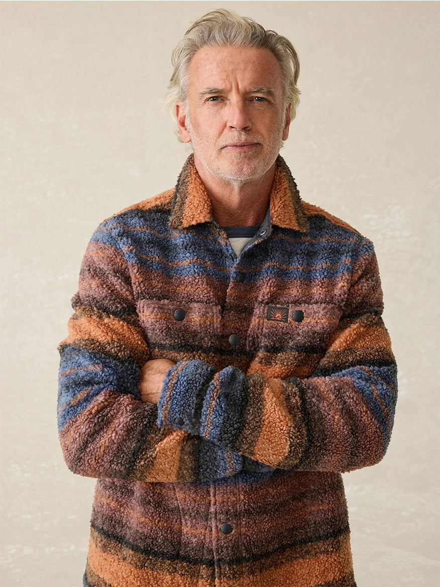 An older man with grey hair and a beard stands with arms crossed, wearing the rugged Faherty Brand Range Fleece CPO in Autumn Horizon Stripe, crafted from a knit comfort wool-blend.