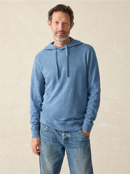 A man wearing a Faherty Brand Sunwashed Slub Hoodie in Aqua Ocean, crafted from American-grown cotton, and jeans stands against a neutral background.