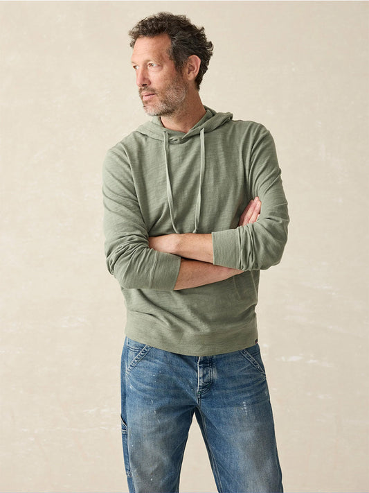 A man with a beard and curly hair stands confidently with his arms crossed, wearing the Faherty Brand Sunwashed Slub Hoodie in Clover Creek, crafted from American-grown cotton, and blue jeans. The relaxed fit of his attire complements the neutral background perfectly.