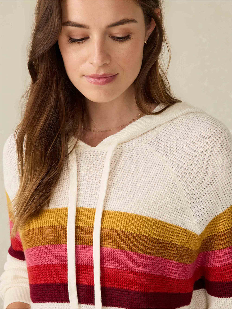 A person wearing the Faherty Brand Throwback Hoodie in Golden Trek Stripe, featuring horizontal stripes in white, yellow, pink, and red, gazes down with a slight smile. This hooded sweater is crafted from organic cotton for ultimate comfort.