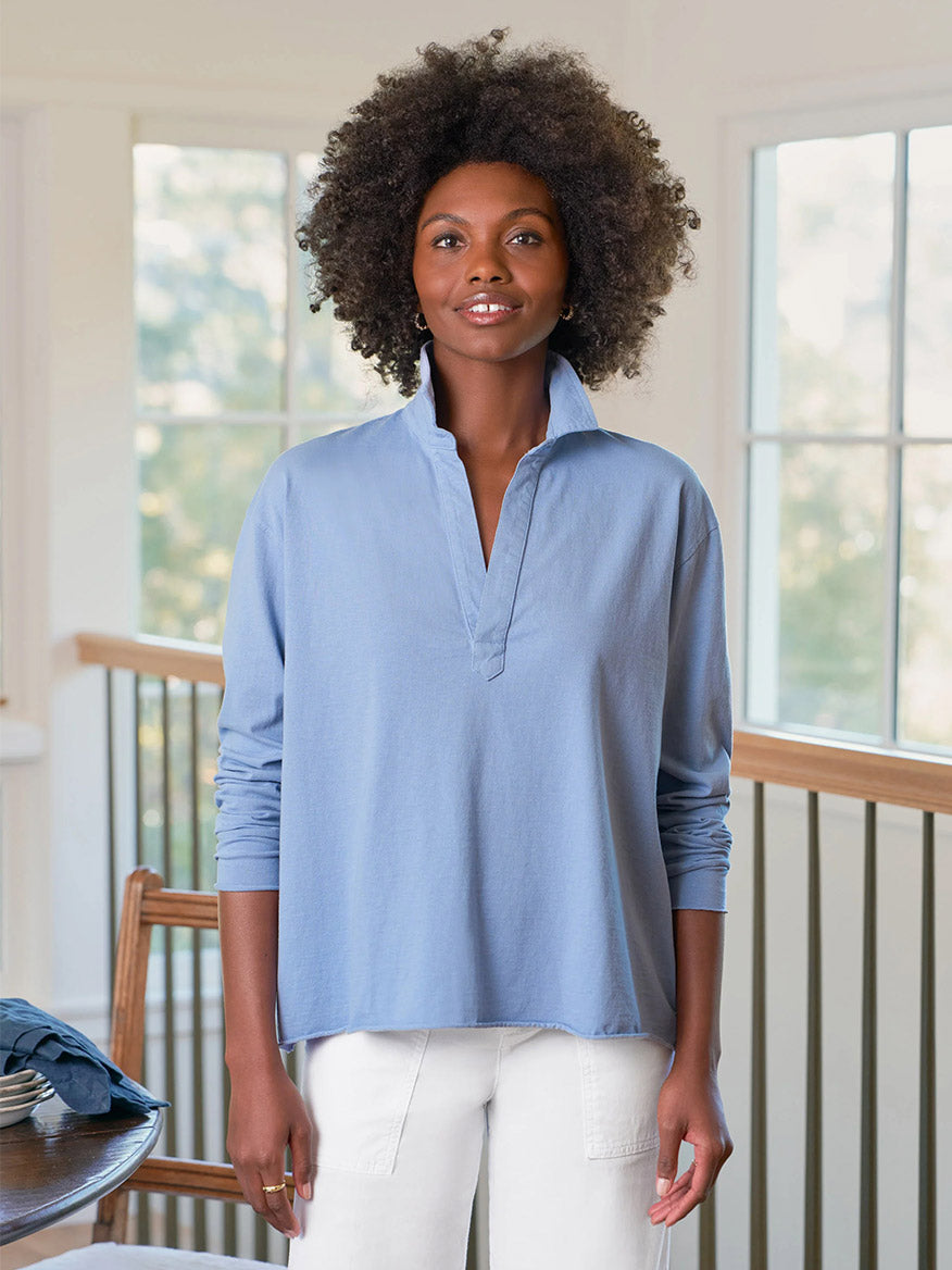 A woman stands indoors by large windows and a wooden railing, wearing white cropped raw hem pants and the Frank & Eileen Patrick Popover Henley in Fog Vintage Jersey. Made in California.