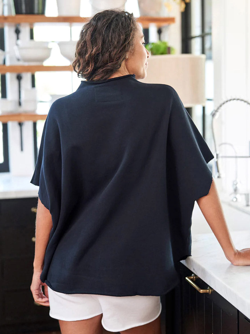 A woman standing in a kitchen, viewed from behind, wearing a Frank & Eileen Audrey Funnel Neck Capelet in British Royal Navy Triple Fleece and white shorts.