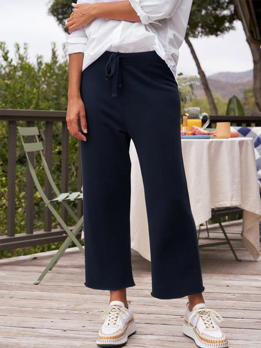 A person standing on a wooden deck wearing white Frank & Eileen Catherine Favorite Sweatpants in British Royal Navy Triple Fleece and dark wide-leg cropped trousers, with a white shirt tucked in at the front.