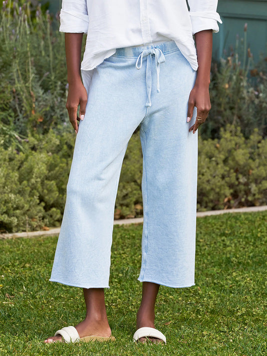 A person wearing a white long-sleeve shirt, Frank & Eileen Catherine Favorite Sweatpant in Mineral Blue Triple Fleece, and white slide sandals stands on grass with greenery in the background.