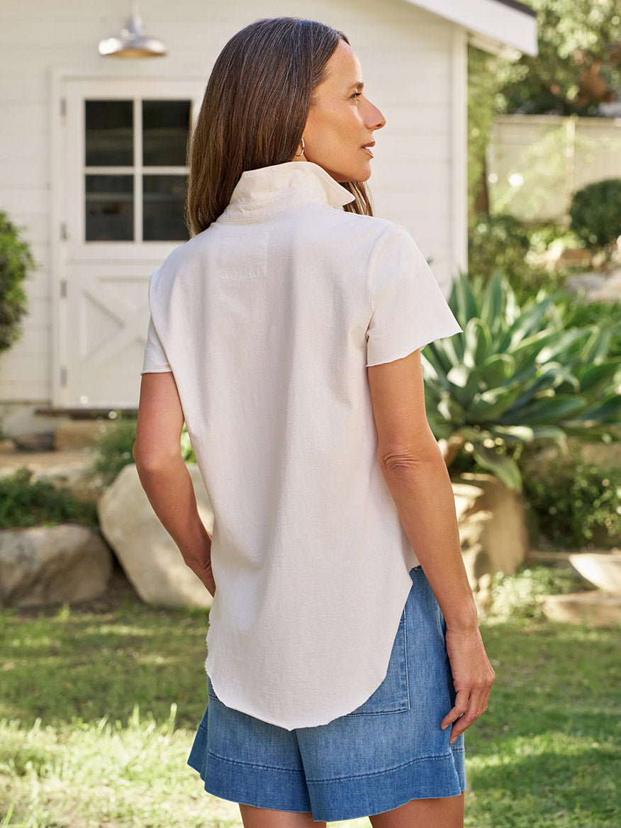 A woman stands outdoors with her back to the camera, wearing the Frank & Eileen Charlotte Perfect Polo in Vintage White Heritage Jersey and denim shorts with a raw hem, against a backdrop of a white building and lush greenery.