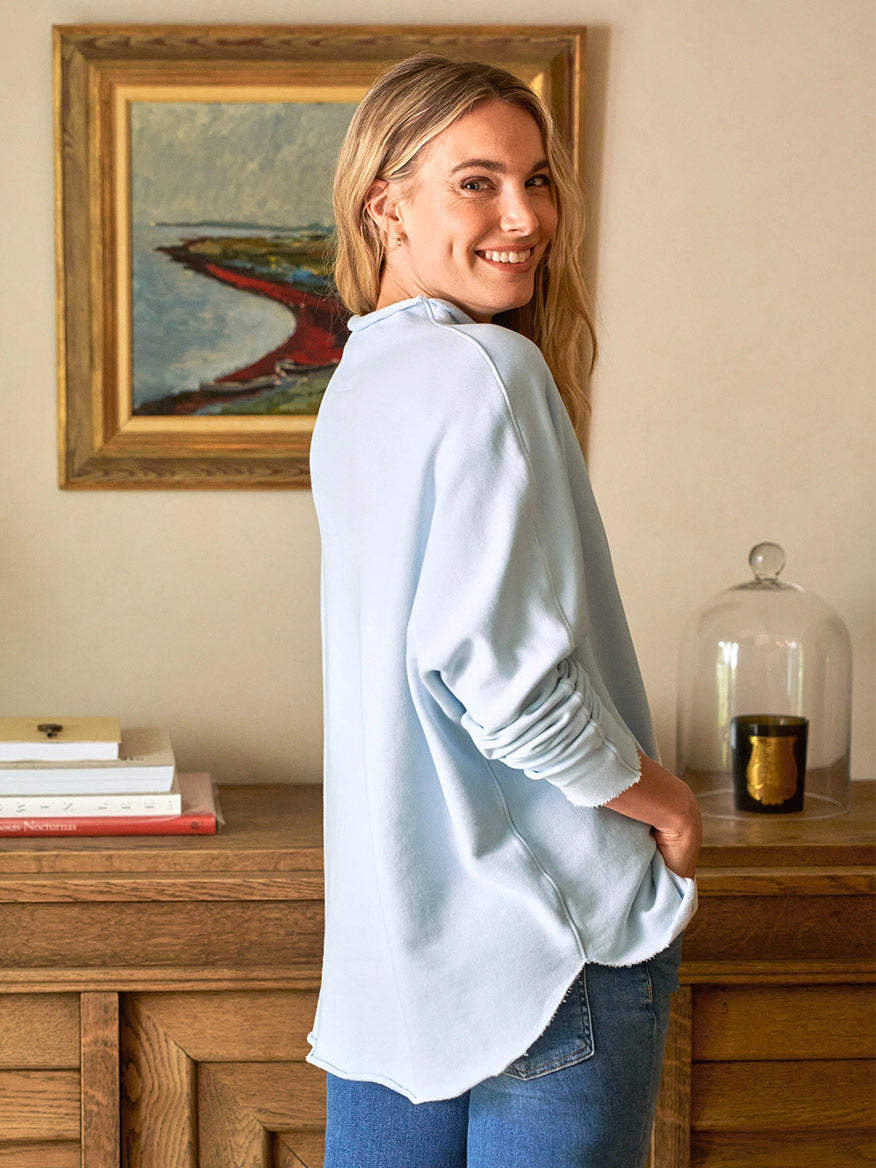 A person in a cozy Frank & Eileen Effie Long-Sleeve Funnel Neck Capelet in Ice Triple Fleece and jeans stands smiling, hands in pockets, amidst a painting and books on a wooden table.