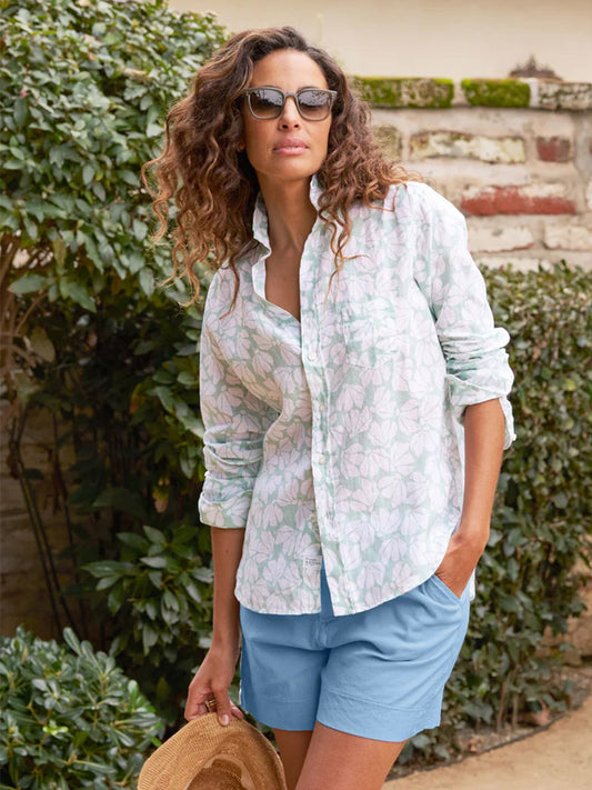 A woman with curly hair stands outdoors, wearing sunglasses, a Frank & Eileen Eileen Relaxed Button-Up Shirt in Mint Floral Classic Linen with signature bust-flattering button placement, and blue shorts, holding a straw hat.