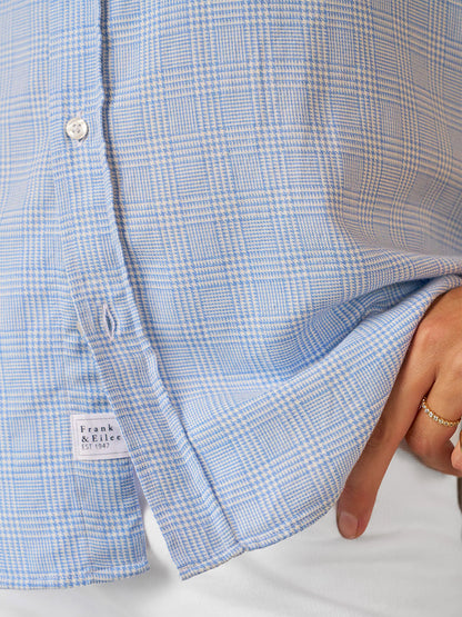 Close-up of a person in the Frank & Eileen Eileen Relaxed Button-Up Shirt in Light Blue Houndstooth Plaid with white pants, holding the hem—capturing true California effortless style.