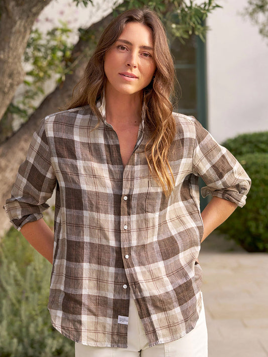 Wearing the Frank & Eileen Eileen Relaxed Button-Up Shirt in Acorn Brown Plaid with rolled sleeves and light-colored pants, a person stands outdoors among lush greenery, embodying an easygoing, casual California vibe.