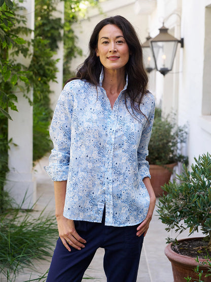 A person in a Frank & Eileen Eileen Relaxed Button-Up Shirt in Blue Floral and dark pants stands outside, hands in pockets, radiating California effortlessness against green foliage and white columns.