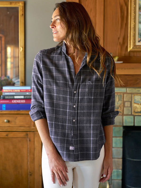 A woman in a Frank & Eileen Eileen Relaxed Button-Up Shirt in Charcoal & Blue Plaid and white pants stands indoors, exuding a casual California vibe as she gazes sideways before a wooden cabinet and tiled fireplace.