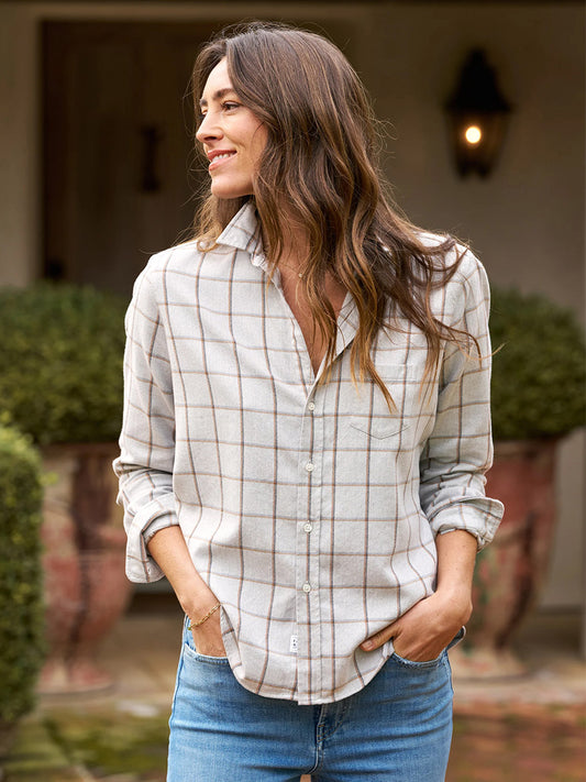 Wearing the Frank & Eileen Eileen Relaxed Button-Up Shirt in Heather Grey with Orange Windowpane paired with jeans, a person stands outdoors with their hands in their pockets, gazing to the side amidst potted plants and near a door.