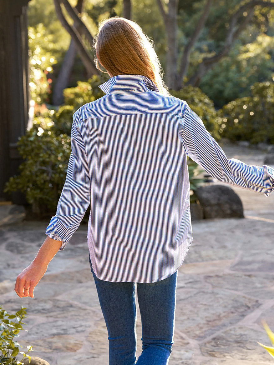 Walking outdoors on a stone path surrounded by greenery, a person with long hair is dressed in a Frank & Eileen Eileen Relaxed Button-Up Shirt in Navy Thin Stripe and jeans with a relaxed fit, exuding California effortlessness.