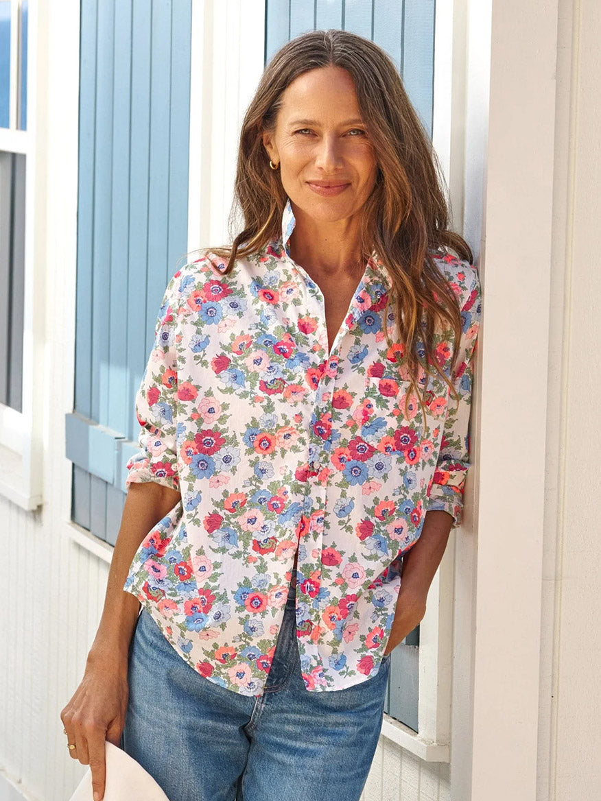 Wearing the Frank & Eileen Eileen Relaxed Button-Up Shirt in Pink and Red with Blue Floral and blue jeans, a woman stands against a white wall with blue shutters, smiling at the camera in effortless, casual California style.