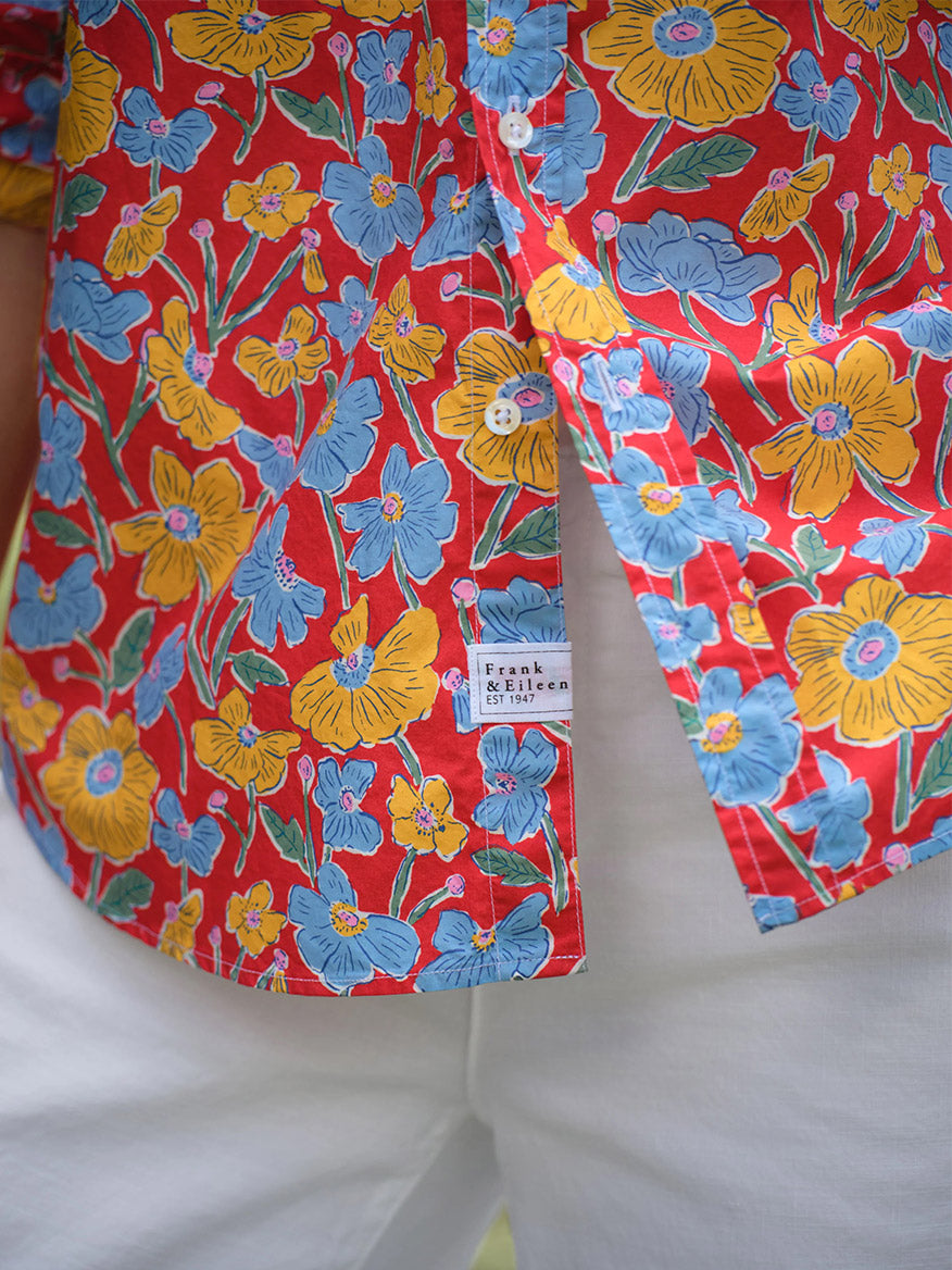 Close-up of a person in white pants and a relaxed, California-style "Frank & Eileen Eileen Relaxed Button-Up Shirt in Red Flowers" featuring blue and yellow florals. The Frank & Eileen label is visible.