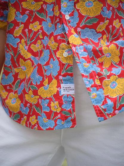 Close-up of a person in white pants and a relaxed, California-style "Frank & Eileen Eileen Relaxed Button-Up Shirt in Red Flowers" featuring blue and yellow florals. The Frank & Eileen label is visible.