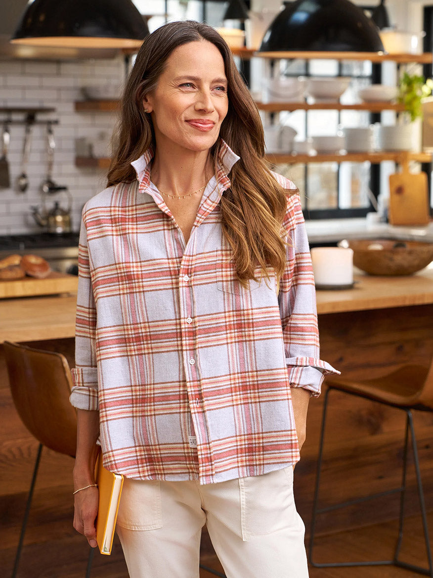 A woman in a Frank & Eileen Eileen Relaxed Button-Up Shirt in Rust & Grey Melange Plaid stands in a kitchen holding a best seller yellow book and smiling.