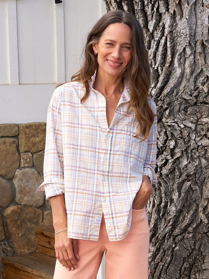 A woman with long hair stands outdoors, smiling, wearing the Frank & Eileen Eileen Relaxed Button-Up Shirt in Sand & Purple Plaid and peach pants with a relaxed fit, her left hand in her pocket. The casual California vibe is evident as she poses near a tree trunk and stone wall in the background.