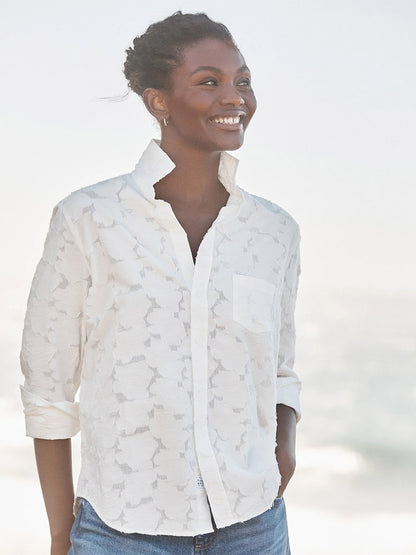 A woman stands by the water, smiling in the Frank & Eileen Eileen Relaxed Button-Up Shirt in Textured White Flowers and blue jeans—capturing effortless California style with its flattering fit and casual vibe.