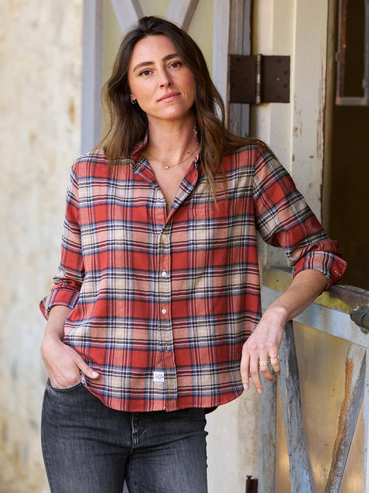 A person leaning against a door frame, effortlessly capturing the California vibe while wearing the Frank & Eileen Eileen Relaxed Button-Up Shirt in Washed Red Plaid and dark jeans, looks at the camera with one hand in their pocket.