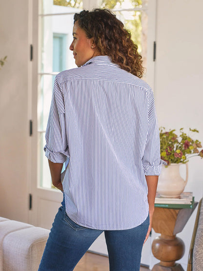A woman stands indoors with her back to the camera, wearing the Frank & Eileen Eileen Relaxed Button-Up Shirt in White & Navy Stripe and jeans. A vase of flowers beside her adds a touch of effortless California style.