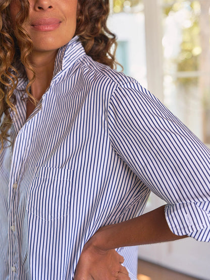 A person with curly hair wears the Frank & Eileen Eileen Relaxed Button-Up Shirt in White & Navy Stripe, sleeves rolled, embodying California effortless style and a relaxed fit indoors, standing with one hand on their hip.