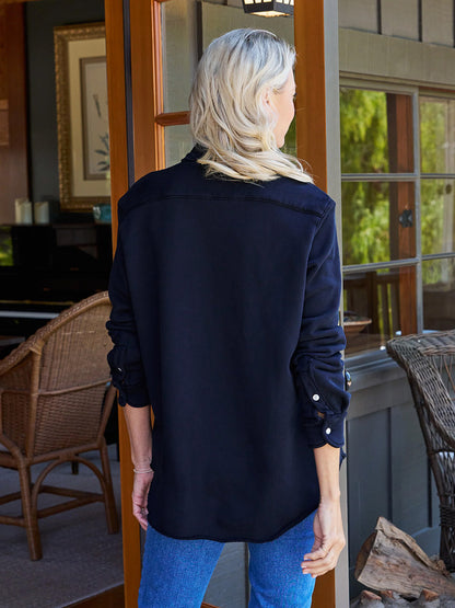 A person with long blonde hair, wearing a Frank & Eileen Eileen Sweatshirt Button-Up in British Royal Navy and relaxed-fit blue jeans, stands with their back to the camera, facing an open door leading to a room with wicker furniture.