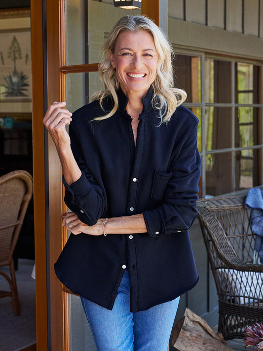 A woman with blonde hair, wearing a Frank & Eileen Eileen Sweatshirt Button-Up in British Royal Navy and blue jeans, stands in an open doorway, smiling. The relaxed fit of her outfit complements the casual California vibe. A wicker chair and a wooden interior setting are visible in the background.