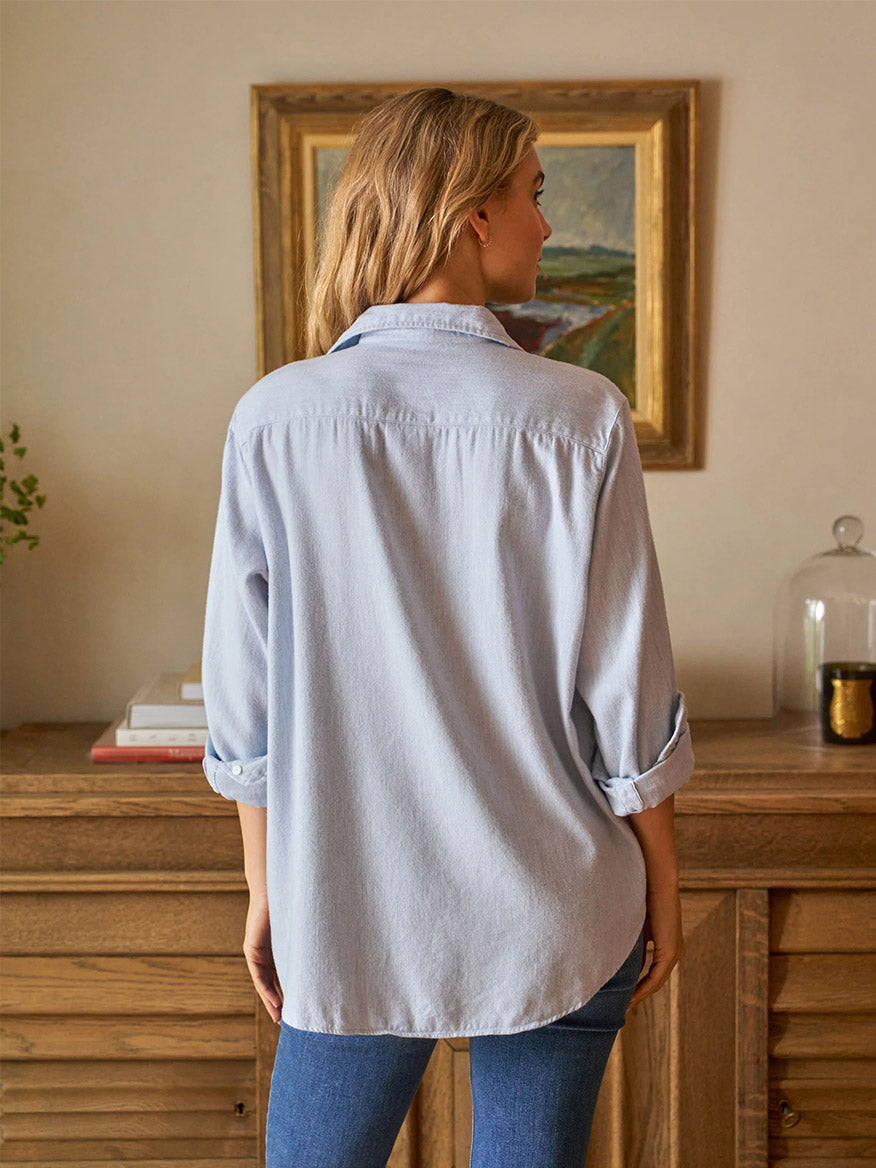 A woman with long hair, embodying California effortlessness, wears a Frank & Eileen Eileen Relaxed Button-Up Shirt in Gray Blue and jeans, standing with her back to the camera in a room decorated with artwork and decor atop a wooden dresser.