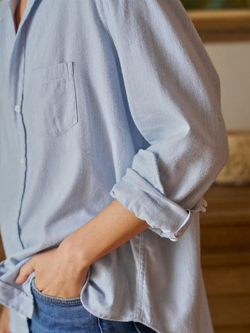 A person exudes California effortlessness, wearing a Frank & Eileen Eileen Relaxed Button-Up Shirt in Gray Blue with bust-flattering buttons and rolled-up sleeves, paired with relaxed-fit blue jeans, standing with one hand in their pocket.