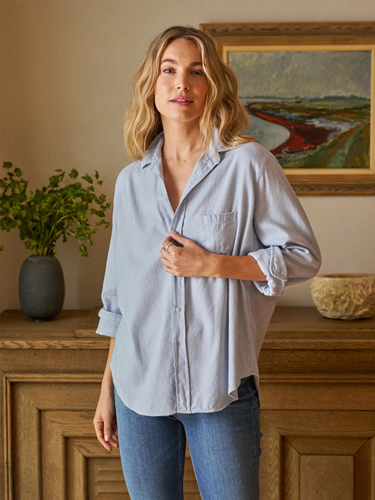 Indoors, a long-haired person exudes California effortlessness in the Frank & Eileen Eileen Relaxed Button-Up Shirt in Gray Blue with bust-flattering buttons, paired with jeans. A painting and plant decorate the wooden background.