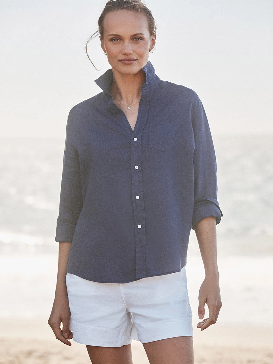 A woman stands on the beach, wearing the Frank & Eileen Eileen Relaxed Button-Up Shirt in Navy Washed Linen and white shorts, embodying casual California style with the ocean behind her.