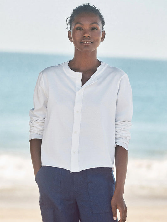 On the beach with the ocean and sky behind, a person wears blue pants, a white button-up shirt, and the Frank & Eileen Jackie T-Shirt Cardigan in White Vintage Jersey in a cropped fit.