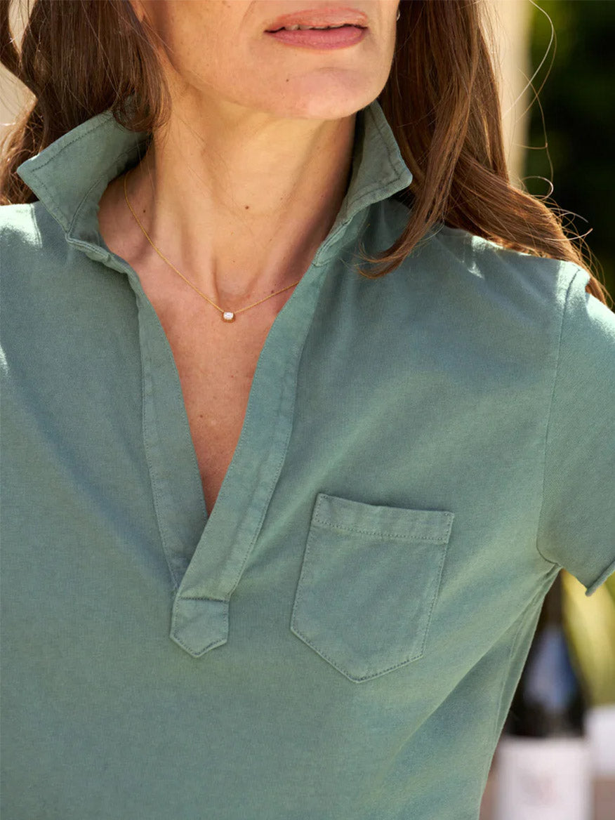 A person wearing a Frank & Eileen Lauren Polo Dress in Rosemary with an open placket and a single chest pocket stands in an outdoor setting. The relaxed fit of this versatile essential provides both comfort and style, complemented by a delicate gold necklace with a small pendant visible around their neck.