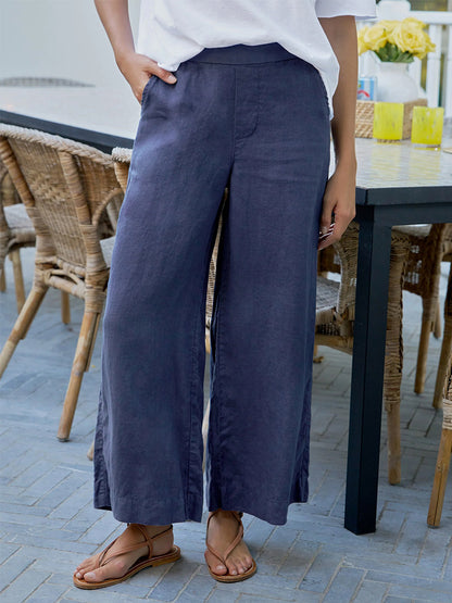 Someone in Frank & Eileen Maisie Wide-Leg Pull-On Pant in Navy Washed Linen, a white top, and brown sandals stands by a black outdoor table with wicker chairs.