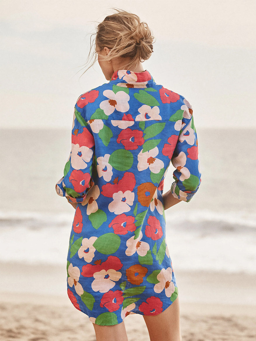 A woman stands on the beach facing the ocean, wearing the Frank & Eileen Mary Classic Shirtdress in Bright Blue Floral—a true wardrobe must-have.