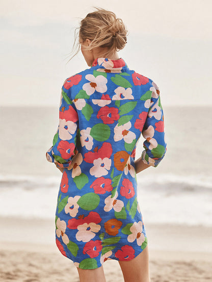 A woman stands on the beach facing the ocean, wearing the Frank & Eileen Mary Classic Shirtdress in Bright Blue Floral—a true wardrobe must-have.