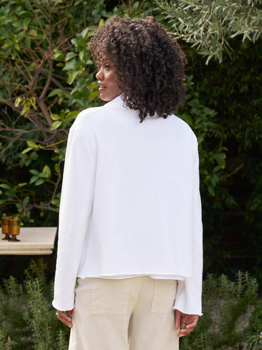 Woman with curly hair wearing a Frank & Eileen Mini Belfast Crop Peacoat in White Triple Fleece shirt and beige pants, standing in a garden, viewed from the back.