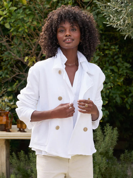 A woman with curly hair wearing a Frank & Eileen Mini Belfast Crop Peacoat in White Triple Fleece and beige pants stands in a garden, buttoning her jacket.