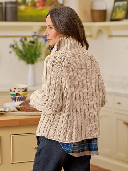 A woman stands in a kitchen facing away, wearing the Frank & Eileen Mini Cotswolds Cropped Cable Knit Cardigan in Sand over a blue plaid shirt. A vase of flowers and dishes sit on the counter.